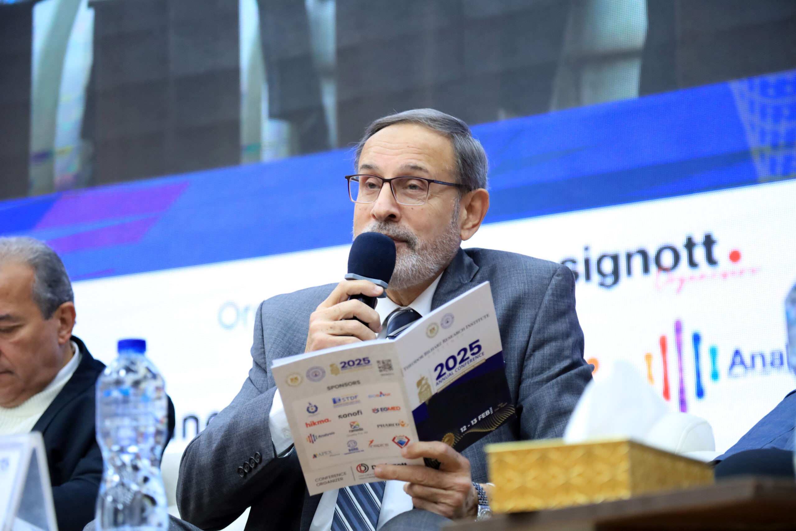 Chairperson smiling while speaking into a microphone and holding a Designott brochure during a conference