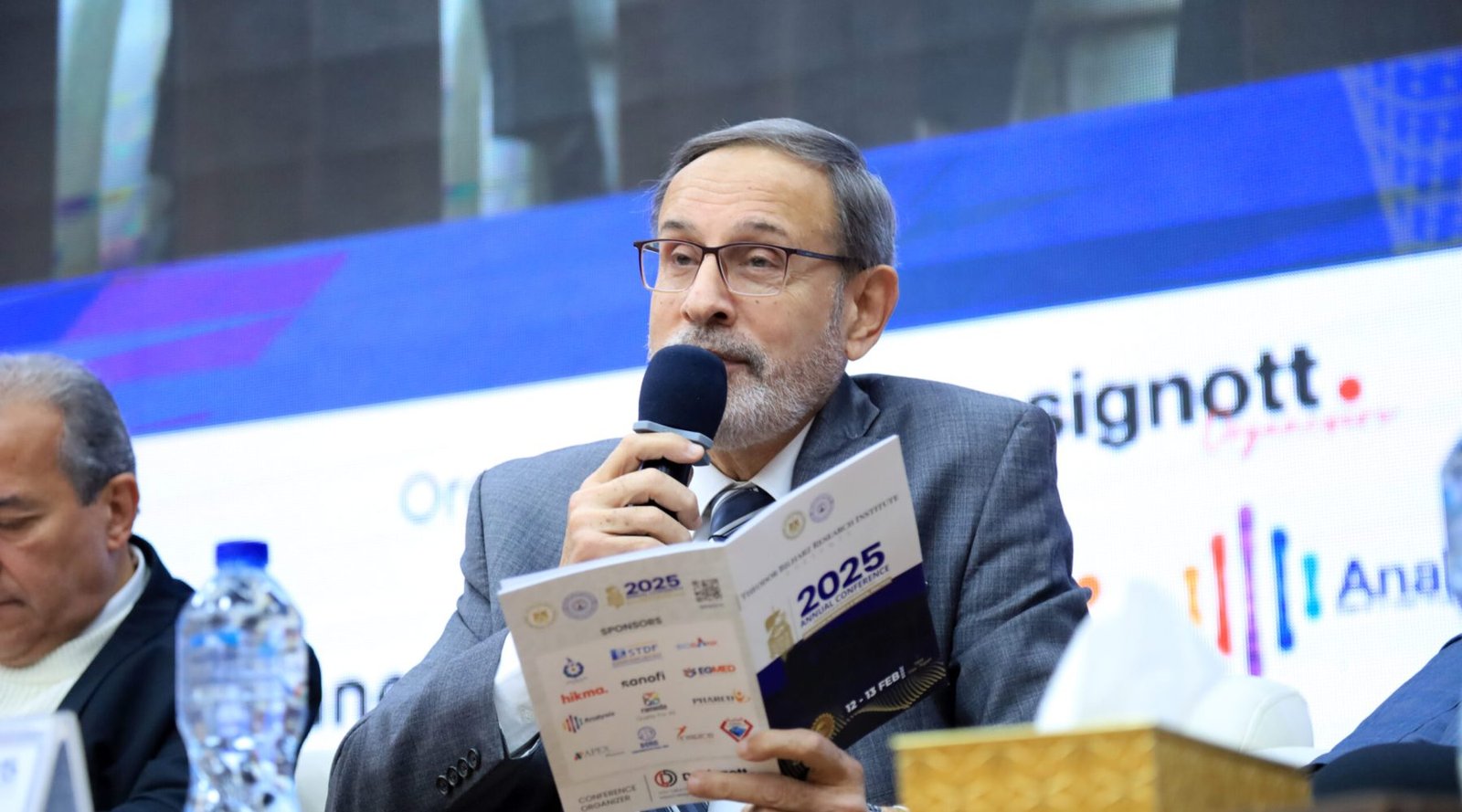Chairperson smiling while speaking into a microphone and holding a Designott brochure during a conference